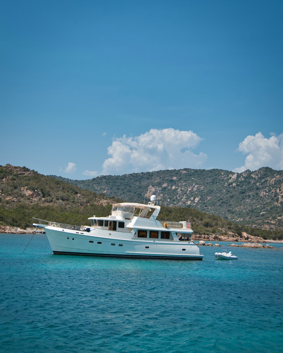 A trawler-style motor yacht at anchor in a turquoise bay below wooded hills.
